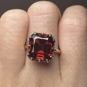 May include: A gold ring featuring a large, rectangular, deep red gemstone. The gemstone is set in a dark, ornate bezel. The ring is shown on a person's finger.