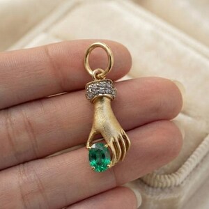 May include: A gold pendant shaped like a hand holding a small oval-cut green gemstone. The wrist area of the hand is adorned with a band of tiny clear stones, and a circular gold bail is attached to the top for hanging on a chain or necklace.
