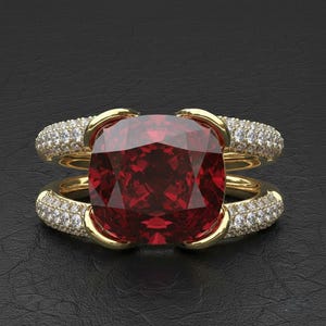 May include: A gold ring featuring a large, cushion-cut, deep red gemstone. The ring has a double band design, with each band adorned with small, sparkling white gemstones. The setting is gold, and the ring is displayed against a black background.