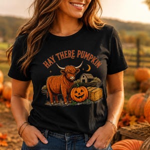 May include: A black short-sleeve t-shirt featuring a graphic of a brown Highland cow standing next to a jack-o'-lantern and a hay bale. Above the cow, the text reads "HAY THERE PUMPKIN" in an orange font, with a small barn and moon visible.