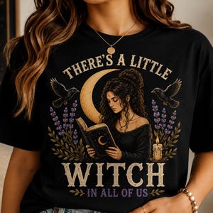 May include: A black t-shirt featuring a graphic print of a person reading a book under a crescent moon. The design includes two black birds, lavender sprigs, and a candle. The text reads: THERE'S A LITTLE WITCH IN ALL OF US in a vintage-style font.