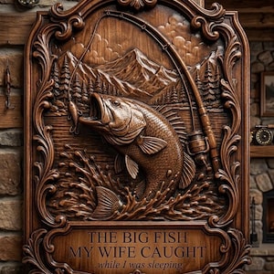 May include: A rectangular wooden wall plaque with ornate carved borders featuring a jumping fish, a fishing rod, and a mountain landscape. The text reads: THE BIG FISH MY WIFE CAUGHT ...while I was sleeping. The wood has a dark brown finish.