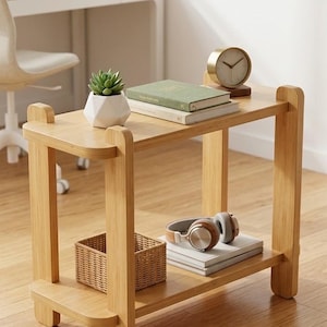 Peut inclure: Une table d'appoint à deux niveaux en bois clair avec des coins arrondis et quatre pieds verticaux. L'étagère supérieure contient un petit pot géométrique blanc pour plante succulente, deux livres empilés et une horloge dorée ronde.