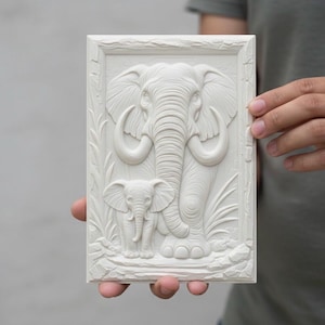 May include: An off-white rectangular relief carving depicting an adult elephant and a baby elephant. The elephants are centrally positioned within a rectangular frame, with carved foliage. The carving is held by a person.