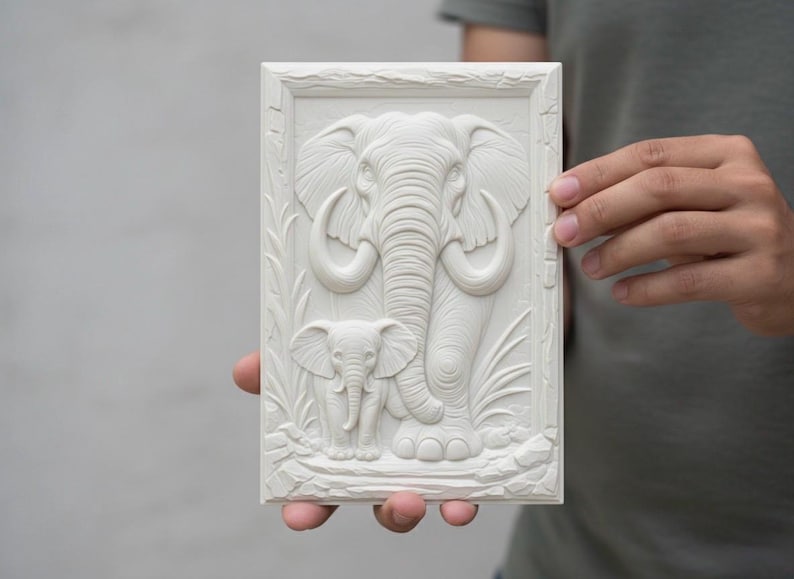 May include: An off-white rectangular relief carving depicting an adult elephant and a baby elephant. The elephants are centrally positioned within a rectangular frame, with carved foliage. The carving is held by a person.
