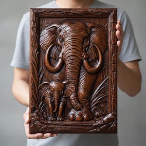 May include: A framed wooden carving depicting an elephant and its calf. The relief sculpture is a warm brown colour, with detailed carvings of the elephant's skin, tusks, and surrounding foliage. The artwork is held by a person.