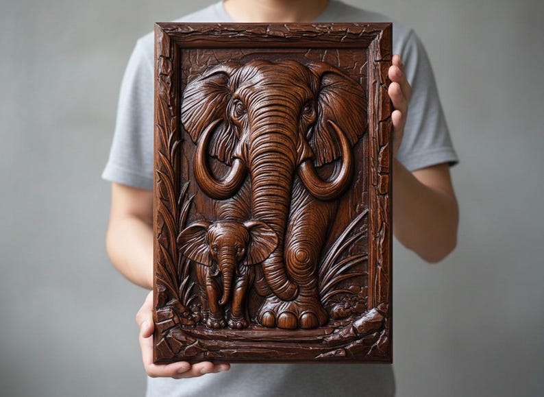 May include: A framed wooden carving depicting an elephant and its calf. The relief sculpture is a warm brown colour, with detailed carvings of the elephant's skin, tusks, and surrounding foliage. The artwork is held by a person.