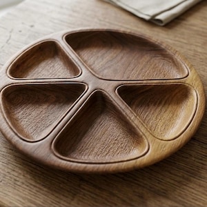 May include: A round, wooden serving tray with five separate compartments. The tray is made of light brown wood and sits on a wooden table. The compartments are triangular and curved, perfect for snacks or appetizers.