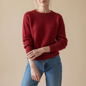 May include: A deep red crew neck jumper with long sleeves. The jumper is made from a soft, textured fabric and is worn with blue jeans. The style is casual and comfortable, suitable for everyday wear.