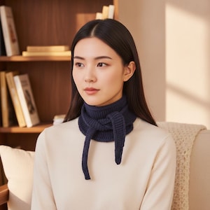 Könnte beinhalten: Ein marineblauer Strickschal wird um den Hals getragen, mit langen, herabhängenden Enden. Der Schal ist aus einem dicken, strukturierten Garn gefertigt. Die Person trägt einen cremefarbenen Pullover. Der Hintergrund umfasst ein Bücherregal aus Holz und ein cremefarbenes Sofa.