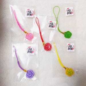 以下が含まれることがあります： カラフルな日本の幸運のお守りストラップのコレクション。各ストラップは、ピンク、赤、緑、黄色、紫など、さまざまな色の織り込まれた結び目デザインが特徴です。各チャームは、お揃いのコードに取り付けられ、日本語のテキストが書かれた小さなカードと一緒にパッケージされています。