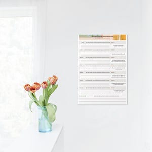 Op de afbeelding: Een witte wandkalender met Franse tekst wordt weergegeven. De kalender heeft een kleurrijke koptekst en een wekelijkse indeling met secties voor elke dag. Een vaas met oranje tulpen staat op een wit oppervlak.