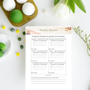 Op de afbeelding: Een weekplanner met de tekst "Weekly Planner" en "Voortgang is belangrijker dan perfectie. Je doet het goed." De planner heeft secties voor elke dag van de week, met ruimte voor notities. Witte en groene decoratieve ballen liggen op een houten dienblad.