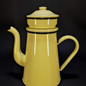 Op de afbeelding: Een lichtgele emaille koffiepot met bijpassend deksel, voorzien van een gebogen tuit en een handvat aan de zijkant. De pot heeft een gladde, glanzende afwerking met dunne zwarte randen langs de opening, de dekselrand en de onderkant.