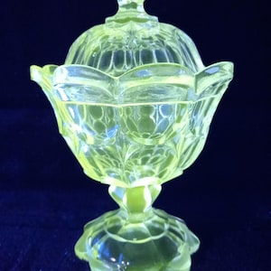 May include: A yellow-green glass candy dish with a lid. The dish has a pedestal base and a decorative, scalloped edge. The lid has a knob on top. The glass has a textured, patterned design.
