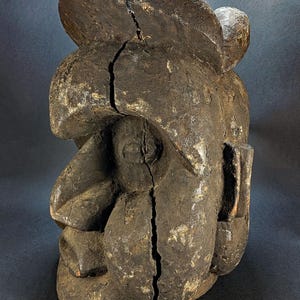 Könnte beinhalten: Eine dunkelbraune, verwitterte Holzskulptur eines stilisierten Kopfes mit einem markanten, abgerundeten Oberteil. Die Oberfläche ist stark strukturiert und weist vertikale Risse auf, die durch die Mitte und über den oberen Teil verlaufen.