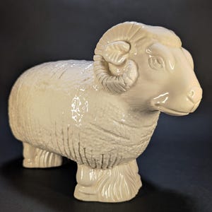 Peut inclure: Une figurine en céramique blanc cassé brillant représentant un bélier debout de profil. La sculpture présente des motifs de laine texturés détaillés sur le corps, des cornes incurvées proéminentes et quatre pattes robustes aux sabots définis.