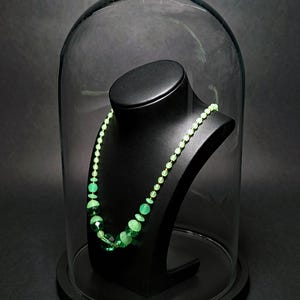Puede incluir: Un collar con cuentas redondas graduadas en varios tonos de verde claro y oscuro translúcido, exhibido en un busto negro bajo una cúpula de cristal. Las cuentas pasan de esferas pequeñas de color verde claro a cuentas facetadas verde oscuro.