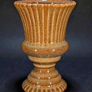 Pode incluir: Um vaso de cerâmica em forma de urna com esmalte castanho salpicado. O recipiente apresenta um rebordo largo e alargado com estrias verticais que se estendem até uma secção média arredondada, estreitando para uma base de pedestal.