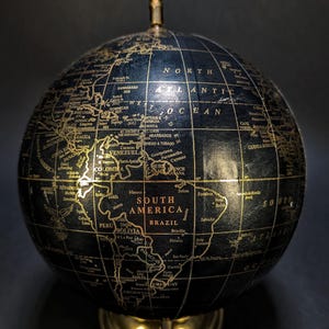 Pode incluir: Um globo esférico com superfície preta, texto e linhas douradas que representam os continentes e oceanos. O globo está montado sobre um suporte metálico de cor dourada com um eixo central e um pequeno remate no topo da esfera.