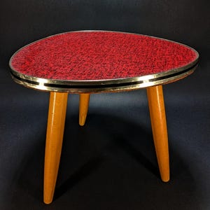 May include: A three-legged side table with a triangular top featuring a textured red and black surface. The tabletop is edged with a metallic band, and the three tapered legs are made of light-coloured wood with a smooth, polished finish.