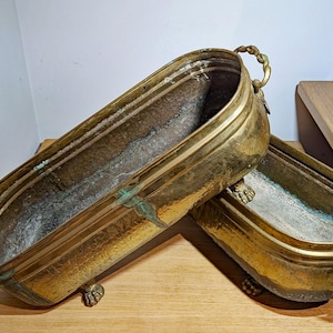 May include: Two oval brass planters with a weathered patina and green oxidation. Each vessel features ornate twisted metal handles on both ends and is supported by four small, detailed claw-style feet. The interior surfaces show signs of age.