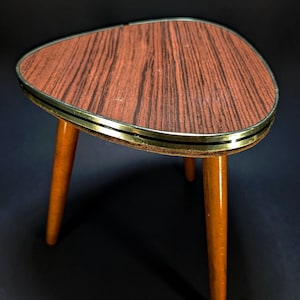 Puede incluir: Una mesa auxiliar pequeña de tres patas con un tablero triangular con patrón de veta de madera marrón oscuro. El borde del tablero está acabado con una banda metálica. Las tres patas cónicas son de madera lisa de color marrón claro.