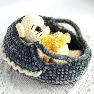 May include: A cream-colored crocheted kitten plush toy with large dark eyes and a pink nose, wearing a yellow crocheted garment. The kitten rests inside a dark gray crocheted basket with a scalloped white trim along the bottom edge.