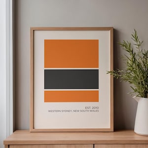 May include: A framed art print with a minimalist design featuring orange and black rectangular blocks. The print includes the text "EST. 2010 WESTERN SYDNEY, NEW SOUTH WALES." A white pitcher with green foliage sits to the right on a wooden surface.