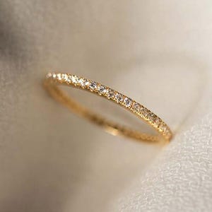 Pode incluir: Um delicado anel de ouro com uma fileira contínua de pequenas pedras preciosas brilhantes. O anel é colocado sobre um fundo branco macio, realçando o seu design elegante. A banda parece fina e refinada, mostrando o brilho das pedras preciosas.