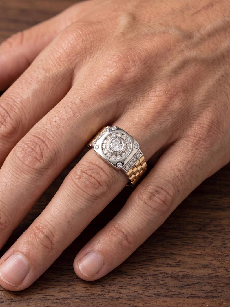 K&ouml;nnte beinhalten: Ein zweifarbiger Ring mit rechteckiger Oberfl&auml;che, der ein zentrales kreisf&ouml;rmiges Design mit kleinen, klaren Steinen aufweist. Der Ring hat ein goldenes Band und eine silberfarbene Oberfl&auml;che mit zus&auml;tzlichen Steinen. Der Ring befindet sich am Finger einer Person.