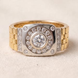 K&ouml;nnte beinhalten: Ein gold- und silberfarbener Ring mit einem Armband im Uhrenstil. Der Ring hat einen zentralen, runden, klaren Edelstein, der von kleineren, klaren Edelsteinen umgeben ist. Das Armband ist goldfarben mit einem Uhren-Glieder-Design. Der Ring liegt auf einer wei&szlig;en Oberfl&auml;che.