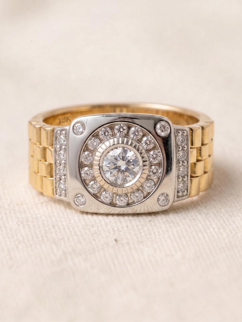 K&ouml;nnte beinhalten: Ein gold- und silberfarbener Ring mit einem Armband im Uhrenstil. Der Ring hat einen zentralen, runden, klaren Edelstein, der von kleineren, klaren Edelsteinen umgeben ist. Das Armband ist goldfarben mit einem Uhren-Glieder-Design. Der Ring liegt auf einer wei&szlig;en Oberfl&auml;che.
