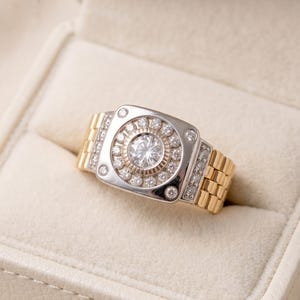 K&ouml;nnte beinhalten: Zweifarbiger Ring mit silberfarbener Oberfl&auml;che und goldfarbenem Band. Die Oberfl&auml;che hat einen zentralen, gro&szlig;en, klaren Stein, der von kleineren Steinen umgeben ist. Das Band hat ein Gliederdesign. Der Ring wird in einer wei&szlig;en Schmuckschatulle pr&auml;sentiert.
