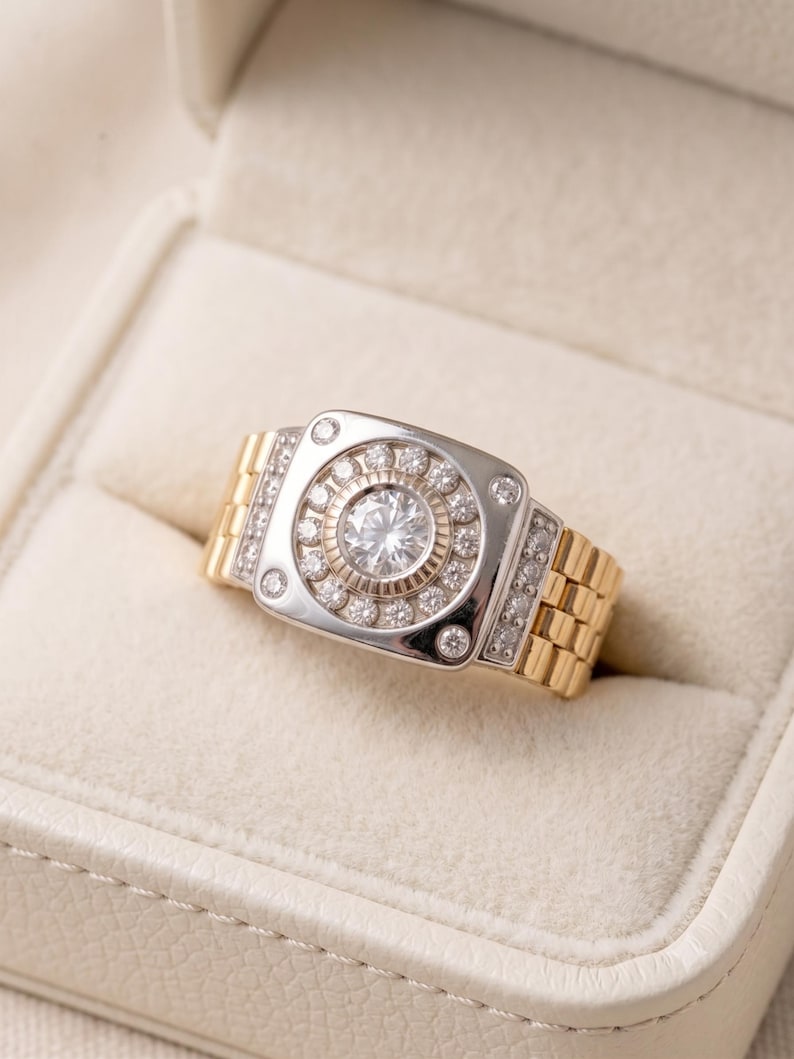 K&ouml;nnte beinhalten: Zweifarbiger Ring mit silberfarbener Oberfl&auml;che und goldfarbenem Band. Die Oberfl&auml;che hat einen zentralen, gro&szlig;en, klaren Stein, der von kleineren Steinen umgeben ist. Das Band hat ein Gliederdesign. Der Ring wird in einer wei&szlig;en Schmuckschatulle pr&auml;sentiert.