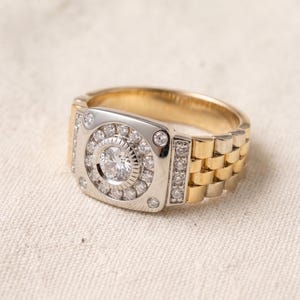 K&ouml;nnte beinhalten: Ein zweifarbiges Gold- und Silberring mit einem gro&szlig;en, runden, klaren Edelstein in der Mitte, umgeben von kleineren Steinen. Der Ring hat ein Design im Uhrenarmband-Stil mit einer rechteckigen Oberfl&auml;che. Das Band ist goldfarben und die Oberfl&auml;che ist silberfarben.