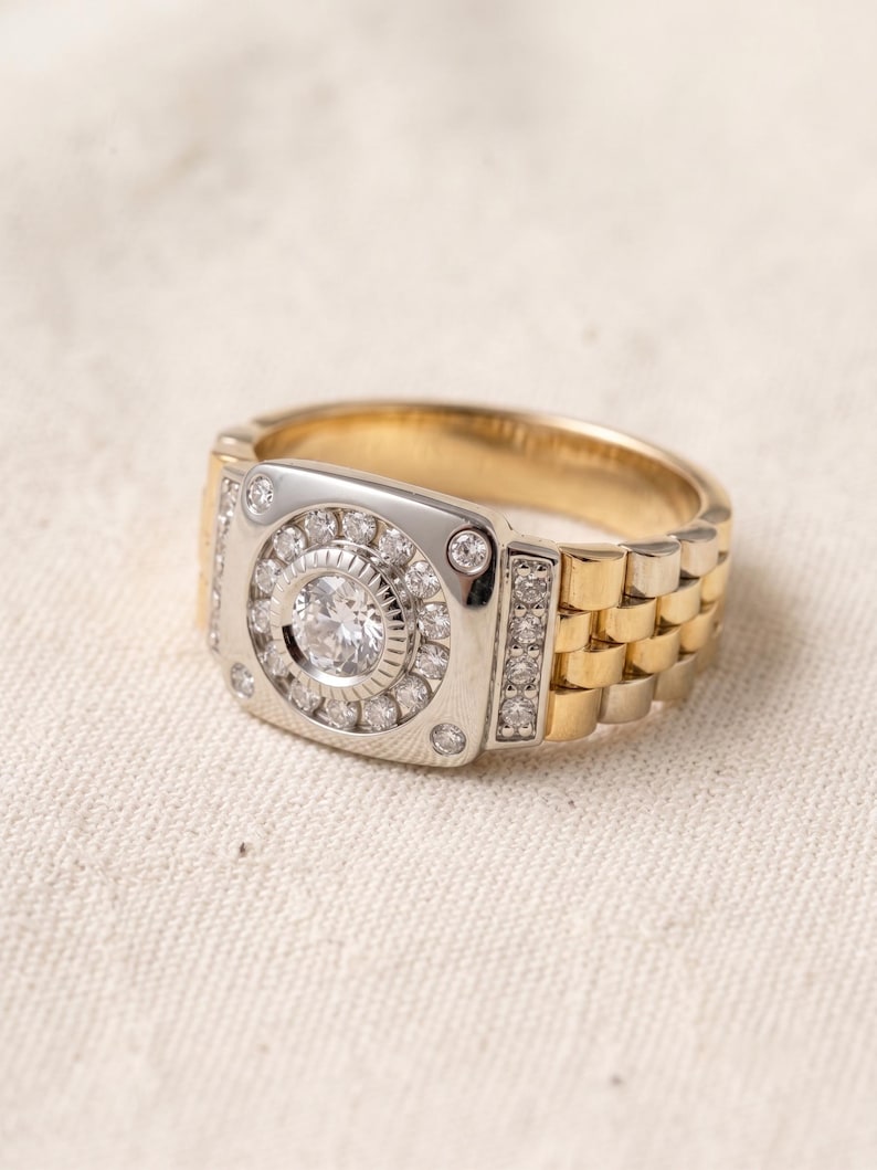 K&ouml;nnte beinhalten: Ein zweifarbiges Gold- und Silberring mit einem gro&szlig;en, runden, klaren Edelstein in der Mitte, umgeben von kleineren Steinen. Der Ring hat ein Design im Uhrenarmband-Stil mit einer rechteckigen Oberfl&auml;che. Das Band ist goldfarben und die Oberfl&auml;che ist silberfarben.