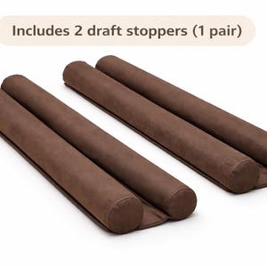 Puede incluir: Cuatro topes de corrientes marrones, dispuestos en dos pares, se muestran sobre un fondo blanco. Los topes son cilíndricos y están hechos de un material suave y texturizado. El texto "Includes 2 draft stoppers (1 pair)" está en la parte superior.