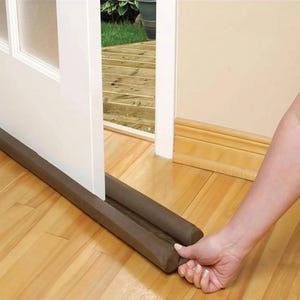 May include: A brown door draft stopper is placed on a hardwood floor. The stopper is positioned at the base of a white door, with a hand holding the end of the stopper. The door is slightly ajar, revealing a view of an outdoor deck.