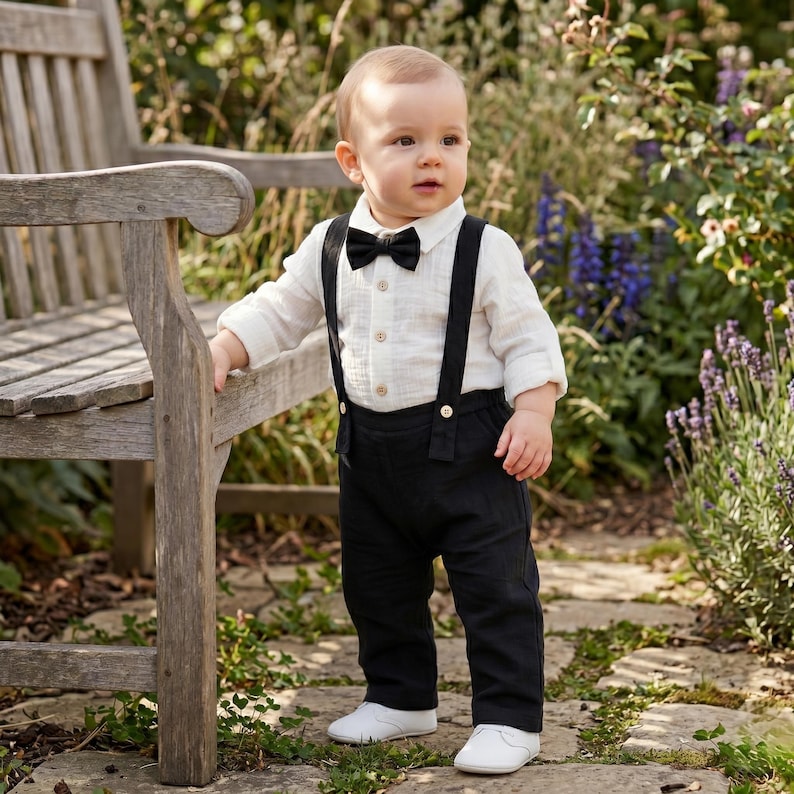 May include: A toddler outfit consisting of a long-sleeved cream-colored button-down shirt, a black bow tie, and black trousers with attached suspenders. The ensemble is completed with plain white shoes, creating a formal look for a young child.