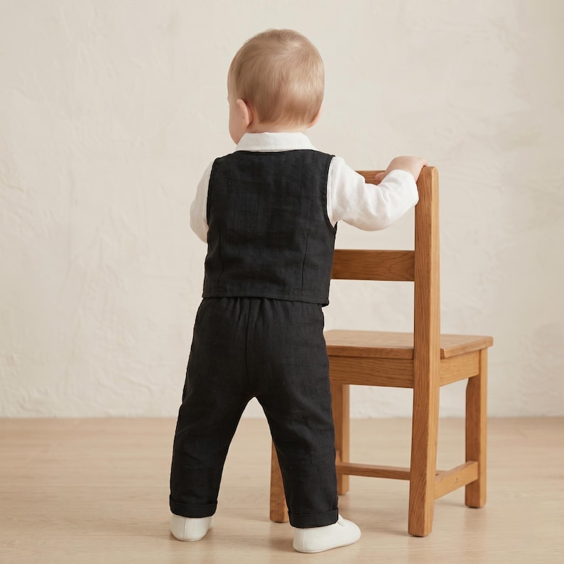 May include: A toddler outfit consisting of a long-sleeved white shirt, a black vest, and matching black trousers. The ensemble is worn by a child standing and holding onto a wooden chair, showcasing the back view of the dark-colored clothing set.