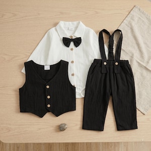 May include: A white long-sleeved button-down shirt with a black bow tie, a black sleeveless vest with three light-colored buttons, and black trousers with attached suspenders. The set features a textured fabric and light-colored button details.
