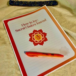 May include: A journal titled "Flow In Joy: Sacral Chakra Journal" with a red and orange border, a red flower graphic, and an orange pen. An incense stick and holder are in the background.