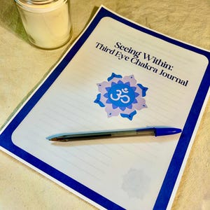 May include: A lit candle in a glass jar next to a journal titled "Seeing Within: Third Eye Chakra Journal." The journal has a blue border and a blue and purple floral design with the symbol "Om." A blue pen rests on the journal.