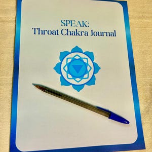 May include: A white journal with a blue border and the text "SPEAK: Throat Chakra Journal" printed in blue. A blue lotus-style chakra symbol is centered below the text. A clear plastic ballpoint pen with a blue cap rests diagonally on the journal.