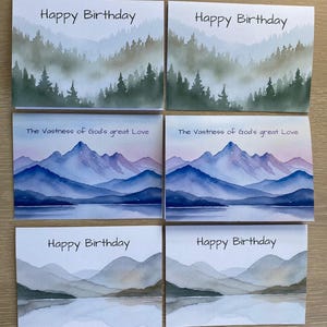 May include: A collection of watercolor greeting cards. Some cards read "Happy Birthday" above a misty forest scene. Other cards display "The Vastness of God's great Love" above a mountain and lake landscape.