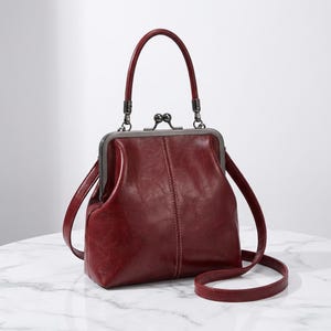May include: A burgundy handbag with a kiss-lock clasp, top handle, and a detachable shoulder strap. The bag has a vintage aesthetic, with visible stitching details and a structured shape. The metal hardware is a dark gunmetal colour.