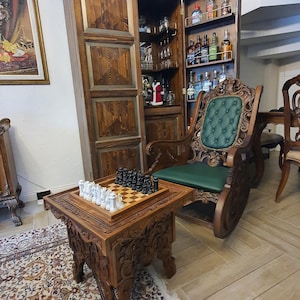 May include: A detailed shot of a chess set on a carved wooden table. The chess pieces are black and white. A dark wooden rocking chair with a green cushion is in the background. A wooden cabinet with bottles is also visible.