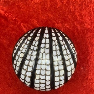 Könnte beinhalten: Runder, dekorativer Gegenstand mit muschelartigem Design. Das Objekt weist abwechselnd schwarze und weiße Abschnitte auf, mit einem Perlmutt-Effekt in den weißen Bereichen. Der Hintergrund ist leuchtend rot.