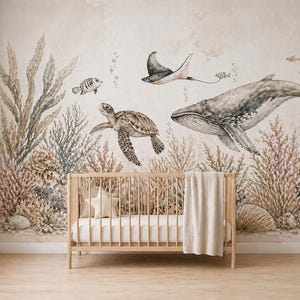 Op de afbeelding: Een kinderkamer met een houten wieg, een beige deken en een stervormig kussen. De muur is versierd met een muurschildering van zeeleven: een walvis, een zeeschildpad en een manta, met waterplanten en schelpen. Lichte, neutrale kleuren.